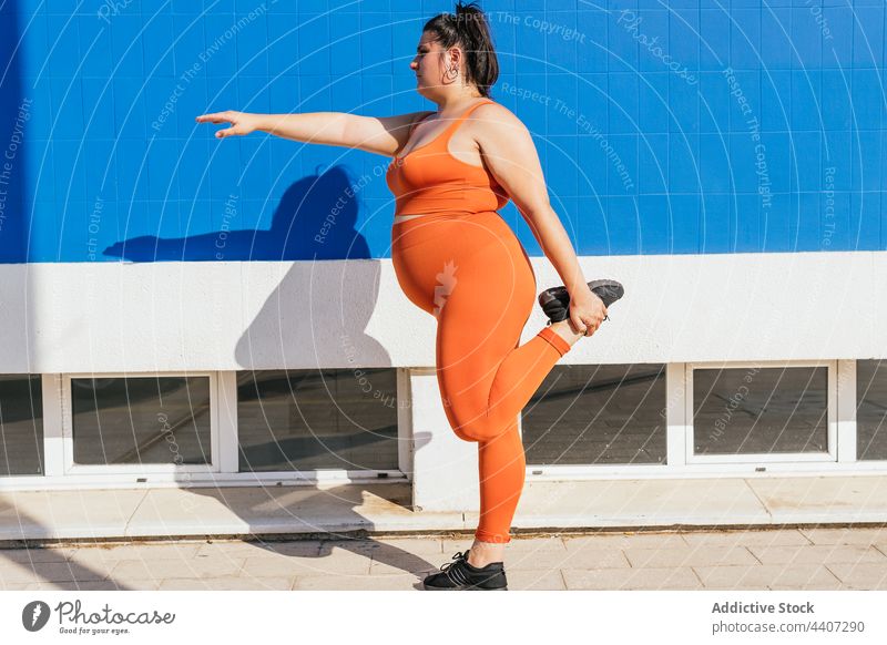 Sportlerin mit kurvigem Körper streckt Bein auf städtischem Bürgersteig Athlet Dehnung Bein angehoben Training Übung mollig Frau Straßenbelag