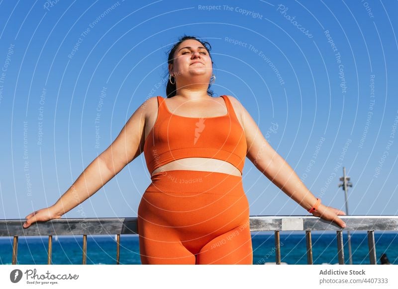 Lächelnde übergewichtige ethnische Sportlerin gegen Zaun und Meer Augen geschlossen verträumt genießen idyllisch Übergröße Stauanlage Blauer Himmel Frau Athlet