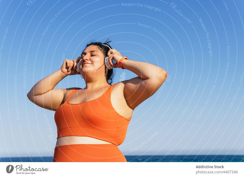 Lächelnde übergewichtige ethnische Sportlerin mit Headset unter blauem Himmel Athlet Kopfhörer zuhören Musik Freizeit Frau Wand benutzend Apparatur Gerät