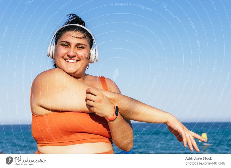 Lächelnde übergewichtige ethnische Sportlerin mit Headset unter blauem Himmel Athlet Kopfhörer zuhören Musik Freizeit Frau Wand benutzend Apparatur Gerät