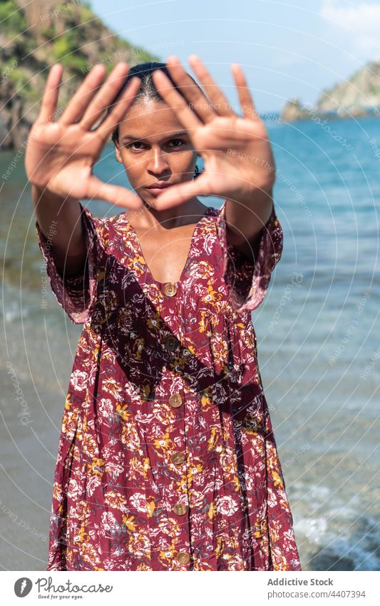Ethnischer Reisender mit Dreiecksgesten am Meeresufer gestikulieren ausdehnen ernst Sonnenkleid Ornament Frau Porträt Starrer Blick geblümt Absicht starren MEER