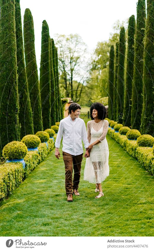Zufriedenes multiethnisches Paar beim Spaziergang durch eine Parkallee Händchenhalten Gasse Partnerschaft Liebe Zusammensein Garten Lächeln rassenübergreifend