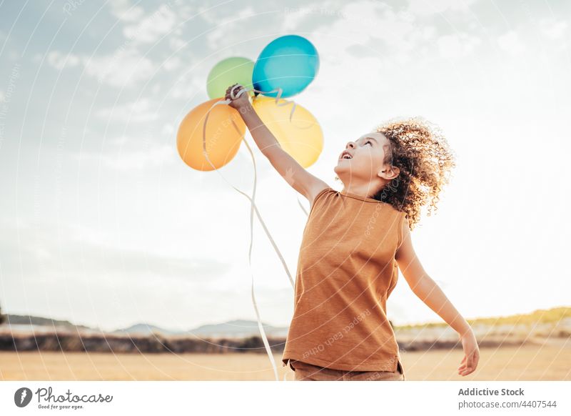 Unbekümmertes ethnisches Kind spielt mit bunten Luftballons im Feld spielen Wind spielerisch sorgenfrei Freiheit Sommer Lächeln heiter Kindheit Glück niedlich