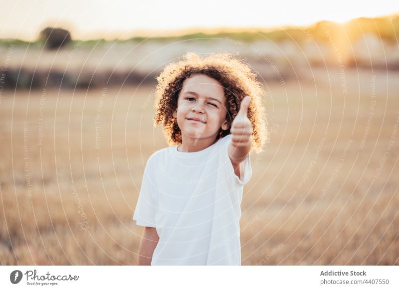 Glückliches ethnisches Kind mit Daumen nach oben im Feld bei Sonnenuntergang mögen gestikulieren Daumen hoch genehmigen weiterempfehlen Zwinkern heiter genießen