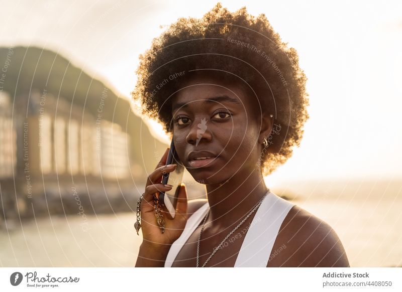 Schwarze Frau, die in der Nähe des Meeres bei Sonnenuntergang mit ihrem Smartphone spricht sprechen Stauanlage MEER reden benutzend Funktelefon Sommer schwarz