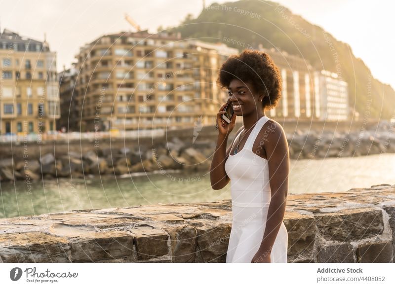 Fröhliche schwarze Frau, die in der Nähe des Meeres bei Sonnenuntergang mit ihrem Smartphone spricht sprechen Stauanlage MEER reden benutzend Funktelefon Sommer
