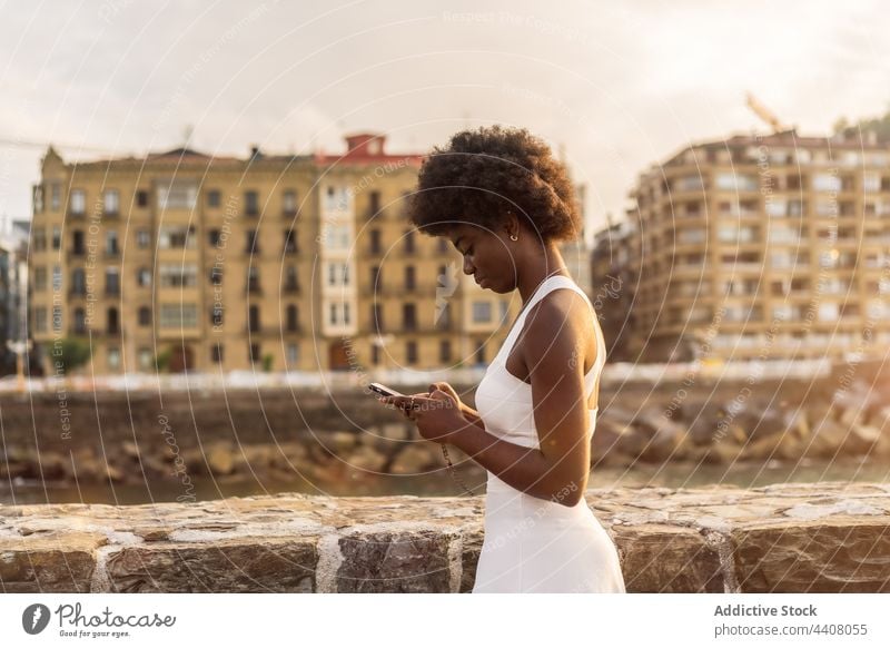Schwarze Frau sendet eine Nachricht auf dem Smartphone in der Nähe des Meeres bei Sonnenuntergang Stauanlage MEER benutzend Browsen Funktelefon Sommer Text