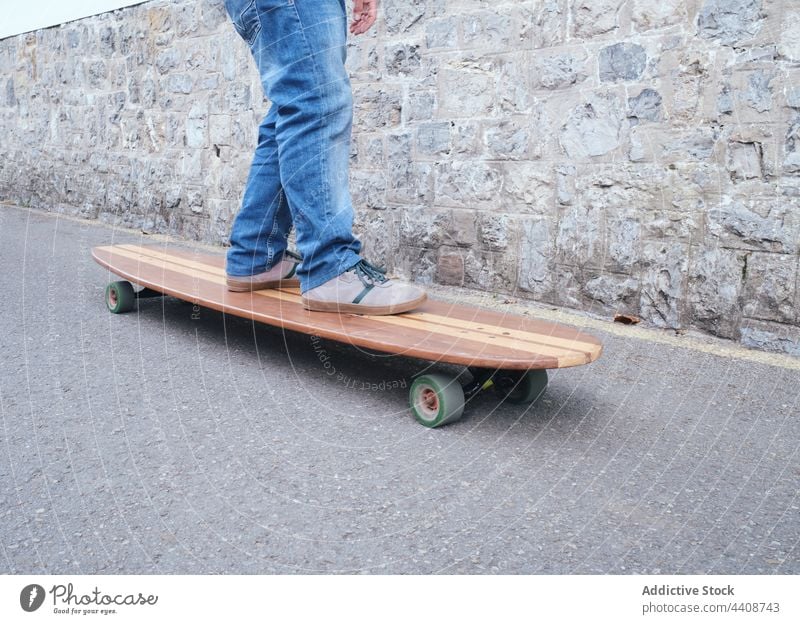 Crop-Skater auf handgefertigtem Longboard auf der Straße Mitfahrgelegenheit aktiv Sport freie Zeit Wochenende Steinwand üben Holzplatte Freiheit Jeanshose