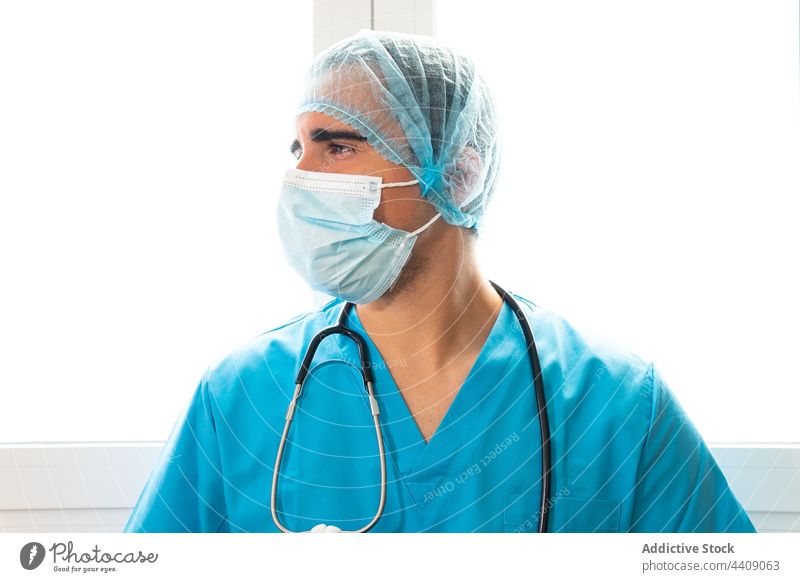 Männlicher Arzt mit Hut und Gesichtsmaske im Krankenhaus stehend Pause Sanitäter Klinik Uniform müde Gesundheitswesen Mann Arbeitstag männlich Job medizinisch