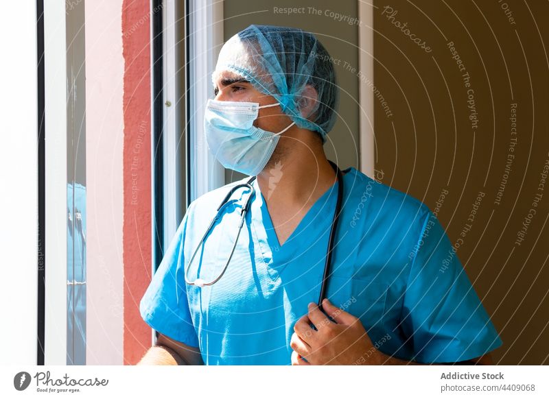 Männlicher Arzt mit Hut und Gesichtsmaske im Krankenhaus stehend Pause Sanitäter Klinik Uniform müde Gesundheitswesen Mann Arbeitstag männlich Job medizinisch