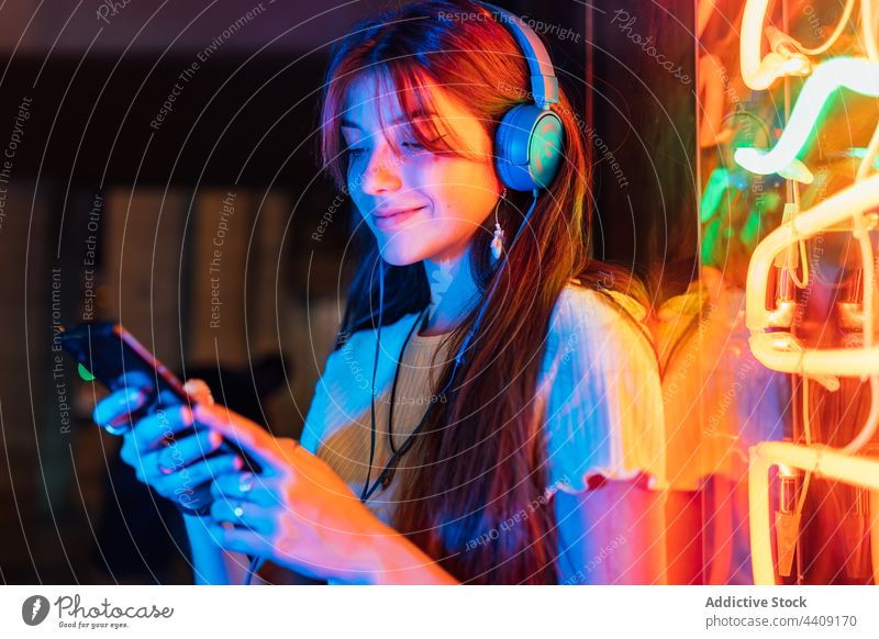 Lächelnde Frau mit Smartphone, die über ein Headset Musik hört Kopfhörer zuhören freie Zeit neonfarbig Stadt benutzend Apparatur Gerät interessiert Internet