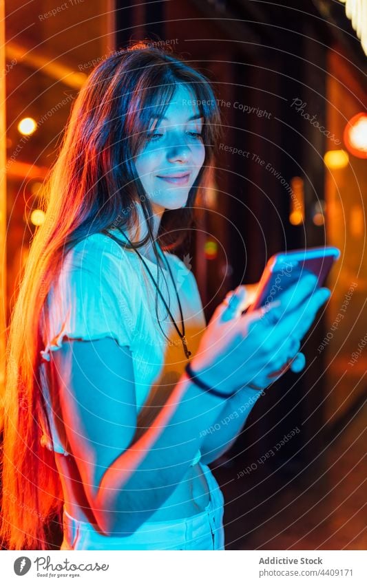 Lächelnde Frau, die in Neonlichtern mit ihrem Smartphone chattet plaudernd neonfarbig interessiert Internet online freie Zeit Stadt benutzend Apparatur Gerät