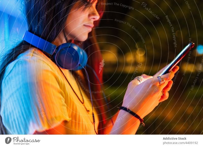 Crop-Frau mit Kopfhörern, die in der abendlichen Stadt mit ihrem Smartphone chattet Textnachrichten Internet Freizeit online Nacht benutzend Apparatur Gerät