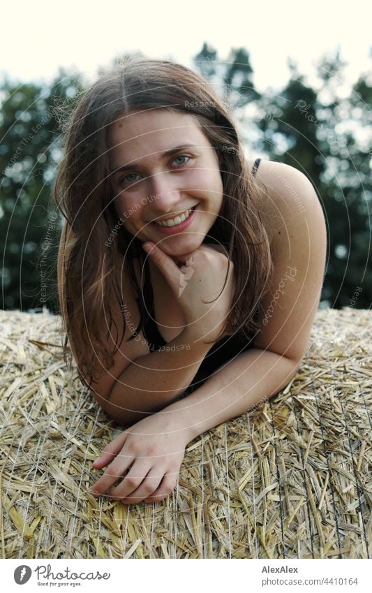 Junge Frau mit Grübchen lächelt und schaut in die Kamera, während sie auf einem Strohballen liegt brünett sportlich 18-30 Jahre Erwachsene langhaarig ästhetisch