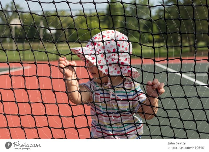 Kleines Mädchen mit Sonnenhut, das sich an einem heißen Tag auf einem Tennisplatz am Netz festhält; das Baby kreuzt und lernt zu laufen - ein Meilenstein in der Entwicklung