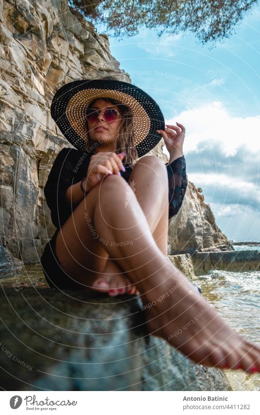 Attraktives Mädchen im schwarzen Kleid, Sonnenbrille und Heuhut am Meer sitzend. Stürmischer Tag am Strand in Kroatien, warmer Sommer 2021. Barfuß Füße im Vordergrund