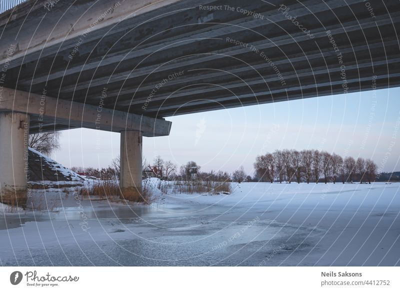 Pfeiler unter der Brücke im Winter auf dem Eis des Flusses. große Transportbrücke in Lettland von unten, Winterabendlicht, klarer Himmel, trockenes gelbes Schilf, schneebedeckter Boden