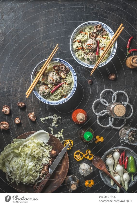 Asiatisches Essen. Schalen mit Nudeln und Shiitake-Pilzen, Chili-Pfeffer, Essstäbchen und Fleischbällchen auf dunklem, rustikalem Hintergrund mit frischen Zutaten der asiatischen Küche und Soßengläsern. Ansicht von oben