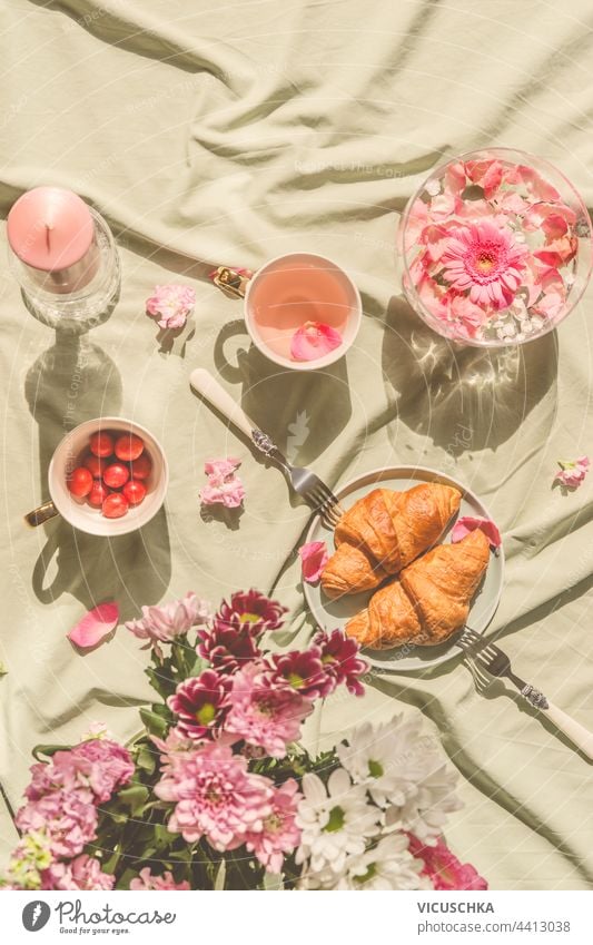 Sonniges ästhetisches Picknick-Frühstück mit Blumen in Kristallvasen, Croissants mit Tee aus Rosenblättern, Blumenstrauß und Kerzen auf Decke. Ansicht von oben. Draußen