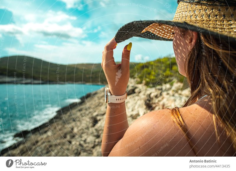 Seitenansicht eines nicht wiederzuerkennenden brünetten Mädchens, das sich im Sommer an ihrem Heuhut festhält. Stehend in der Nähe der Küste des adriatischen Meeres, Konzepte von Reisen, Sommer und Urlaub