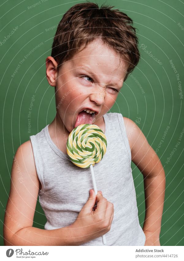 Lustiger Junge leckt süßen Lutscher am Stiel im Studio Kind Lollipop Bonbon lutschen Grimasse Gesicht machen lecker Spaß haben lustig geschmackvoll Leckerbissen