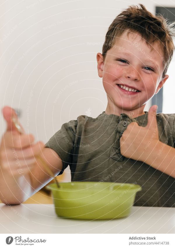 Fröhliches Kind isst Mittagessen und zeigt den Daumen nach oben Daumen hoch mögen gestikulieren gut Lebensmittel Junge Lächeln Glück heiter Küche heimwärts