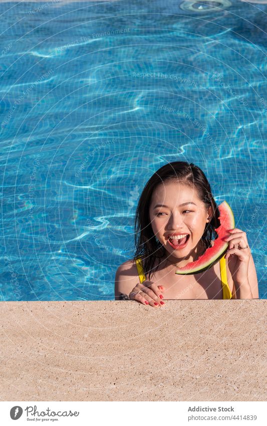 Asiatische Frau hat Spaß mit Wassermelone als Telefon im Schwimmbad Spaß haben Zifferblatt Pool heiter spielerisch Scheibe Sommer ethnisch asiatisch Glück