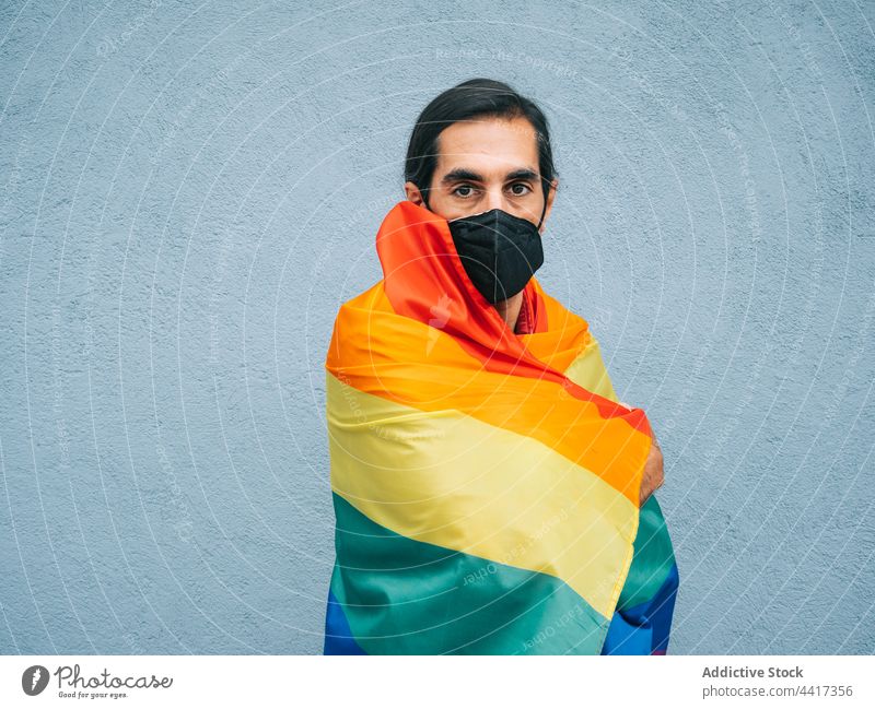 Homosexueller ethnischer Mann mit Maske, eingewickelt in LGBT-Flagge in der Stadt schwul Regenbogen Fahne umhüllen Mundschutz Coronavirus neue Normale Großstadt