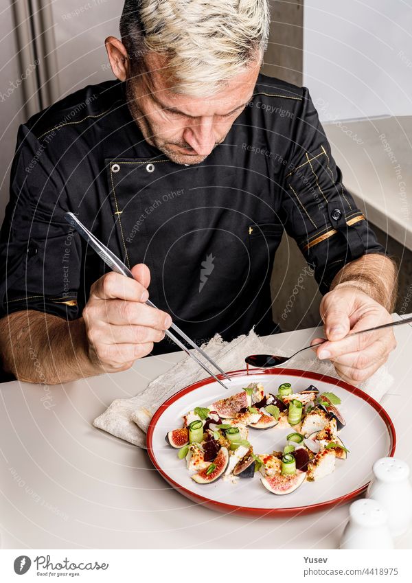 Ein stilvoller Koch im schwarzen Jackett bereitet einen leckeren Salat mit reifen Feigen, Ziegenkäse Chevre, frischen Gurkenröllchen, Roter Bete und Senfblättern mit Tomatensauce zu. Restaurant-Gericht. Leckeres Essen