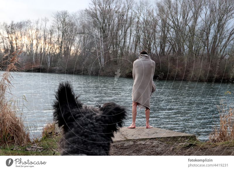 nackter junger Mann steht, in eine Decke gehüllt, an einem winterlichen See und wird von einem Hund beobachtet kalt Herausforderung Winter eisbaden frieren