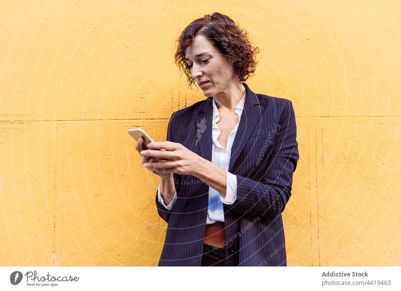Selbstbewusste, gut gekleidete Frau, die in der Nähe einer hellen Wand auf ihrem Smartphone surft elegant formal Stil stilvoll selbstsicher selbstbewusst Glück