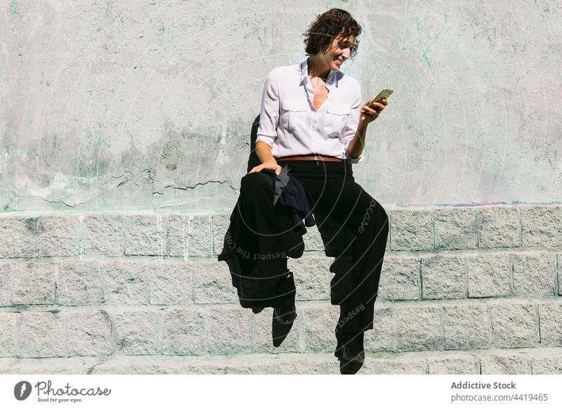 Lächelnde Frau, die auf einem Steinrand sitzt und ihr Smartphone beobachtet benutzend zuschauen online Anschluss Videoanruf positiv formal Gespräch abgelegen