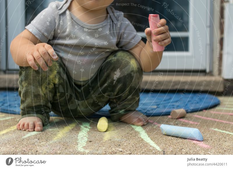 Kleinkind in der Hocke beim Spielen mit Kreide auf der Treppe vor ihrem Haus: Kind trägt eine Hose mit Tarnmuster und ein Oberteil mit Sternenmuster Kniebeuge