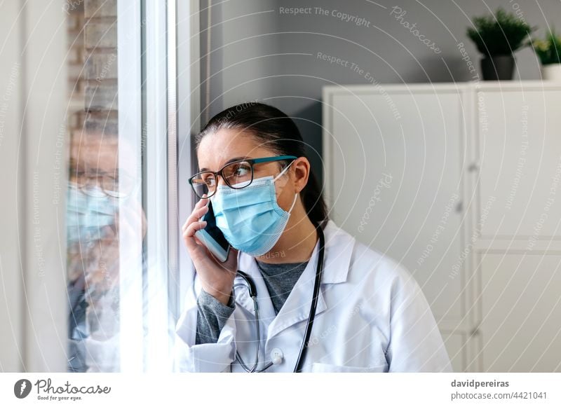 Eine Ärztin telefoniert mit dem Handy und schaut aus dem Fenster Arzt Operationsmaske ernst Coronavirus Ausschau haltend Reflexion & Spiegelung Mobile
