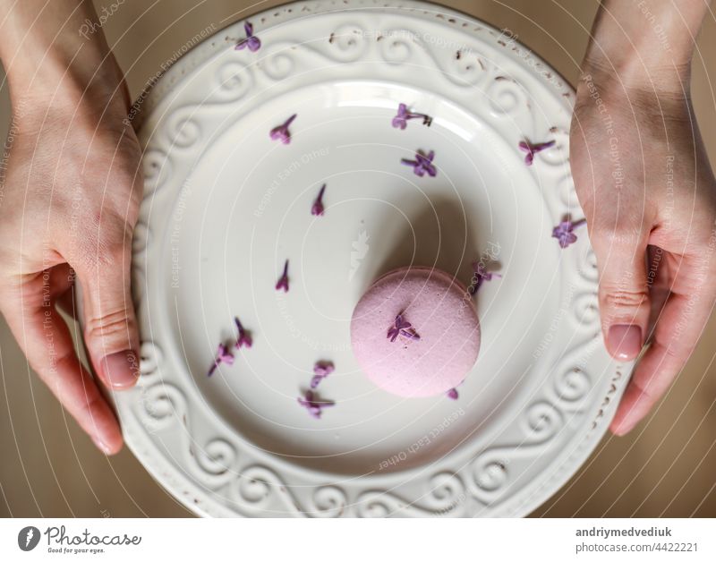 Ausschnitt einer Frau, die einen Teller mit rosa köstlichen französischen Makronen oder Macarons mit lilafarbenen Blumen auf einem hölzernen Hintergrund hält