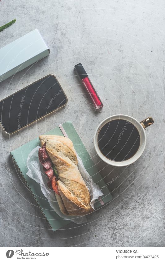 Sandwich-Mittagessen oder Frühstück auf grauem Schreibtisch mit Kaffee, Smartphone und Kosmetik, Ansicht von oben. Belegtes Brot Draufsicht Werkzeug schwarz