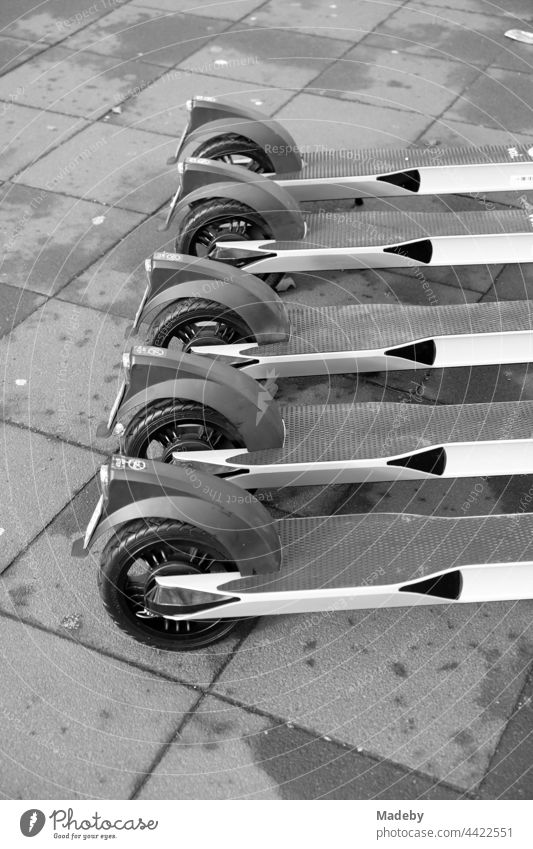 Umstrittene E-Roller zum mieten aufgereiht auf grauem Straßenpflaster in der Innenstadt von Frankfurt am Main in Hessen, fotografiert in neorealistischem Schwarzweiß