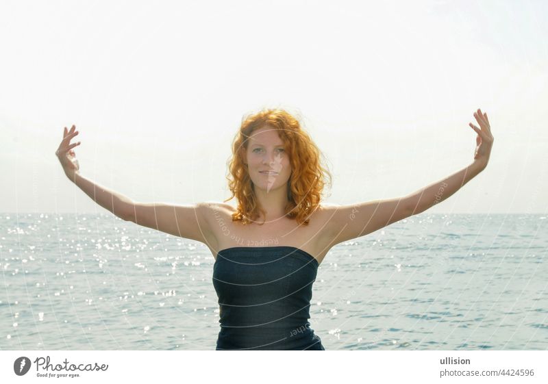Fröhliches Porträt einer jungen, eleganten, rothaarigen, lockigen Frau mit ausgestreckten Armen am Meer am Strand in Italien mit Kopierraum, Platz für Text