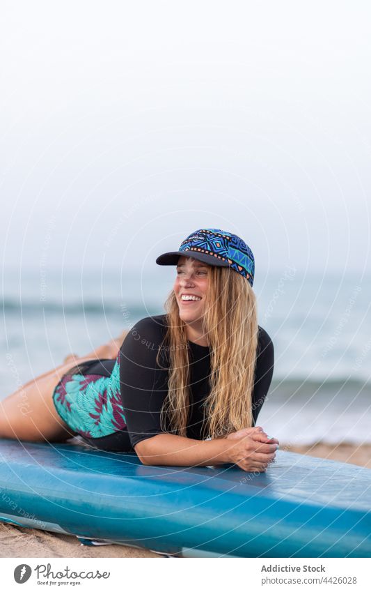 Fröhliche Frau auf SUP-Brett am Strand liegend Surfer Paddelbrett Lügen Zusatzplatine Badeanzug Lächeln Meeresufer MEER Sommer Ufer Urlaub Badebekleidung Hut