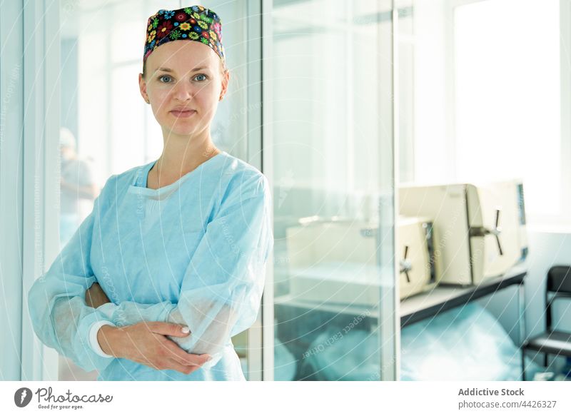 Selbstbewusster Chirurg mit verschränkten Armen in der Klinik selbstsicher die Arme verschränkt Lächeln professionell Uniform Frau Glaswand Porträt Beruf