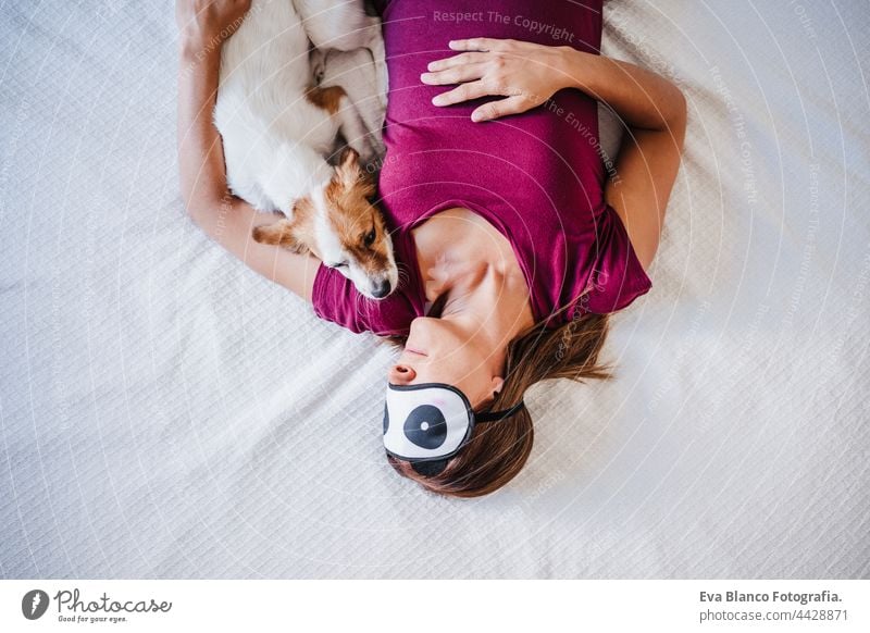 Ansicht von oben entspannte kaukasische Frau zu Hause trägt Panda Augenmaske, während auf dem Bett mit niedlichen Jack Russell Hund ruht. Haustiere, Liebe und Entspannung