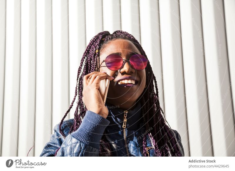 Schwarze Frau mit Zöpfen telefoniert mit einem Smartphone benutzend Afro-Look Stil heiter Zahnfarbenes Lächeln urban trendy Apparatur Geflecht Afroamerikaner
