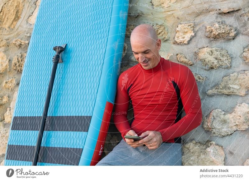 Mann mit Paddelbrett lehnt an der Wand und benutzt ein Mobiltelefon Zusatzplatine Surfer Meeresufer Handy Neoprenanzug Strand männlich Surfbrett Smartphone Sand
