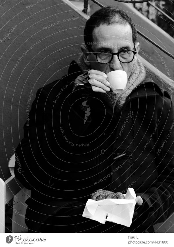 Kalter Kaffee Mann Brille kalt sitzen Winter Schal Bank Jacke Fahrradhandschuhe Croissant trinken Pause Corona Lockdown Sitzbank