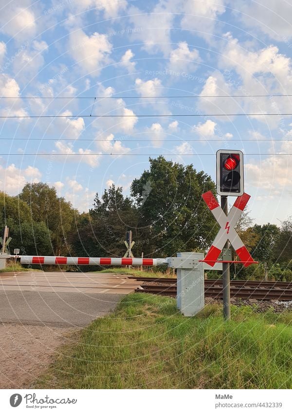 Bahnübergang mit geschlossenen Halbschranken an einem landwirtschaftlichem Weg Andreaskreuz mit Blitz elektrische Oberleitung Stromkasten Lichtanlage rot weiß