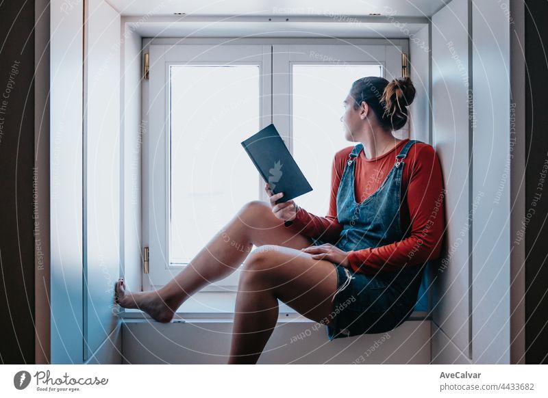Junge Frau, die an einem hellen Tag am Fenster sitzt und ein Buch liest, Konzepte für Reflexion und Selbstfürsorge, Kopierraum lesen aussruhen Appartement