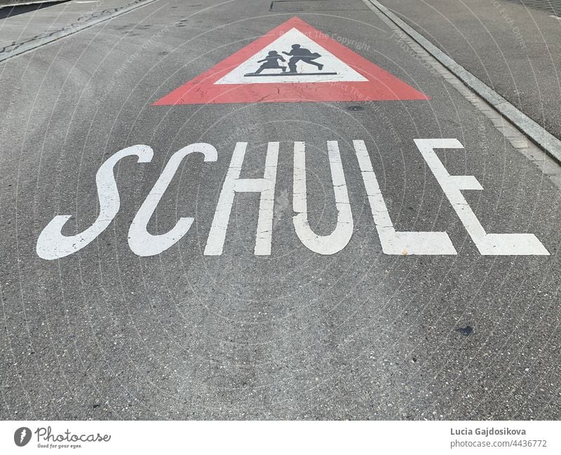 Warnschild für den Schulweg in deutscher Sprache in der Schweiz. Straßenschild ist rotes Dreieck mit laufenden Kindern in weißem Feld und Text Schule, die Schule bedeutet.