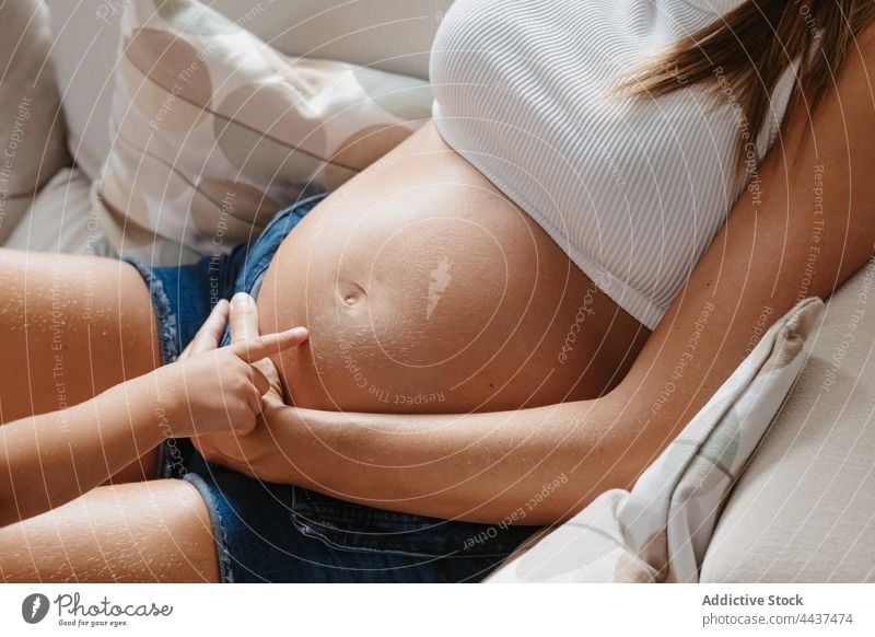 Anonyme schwangere Mutter und Tochter auf dem Sofa interagieren Schwangerschaft Kraulen Zeit verbringen Kindheit Streicheln Mutterschaft Zuneigung Bauch