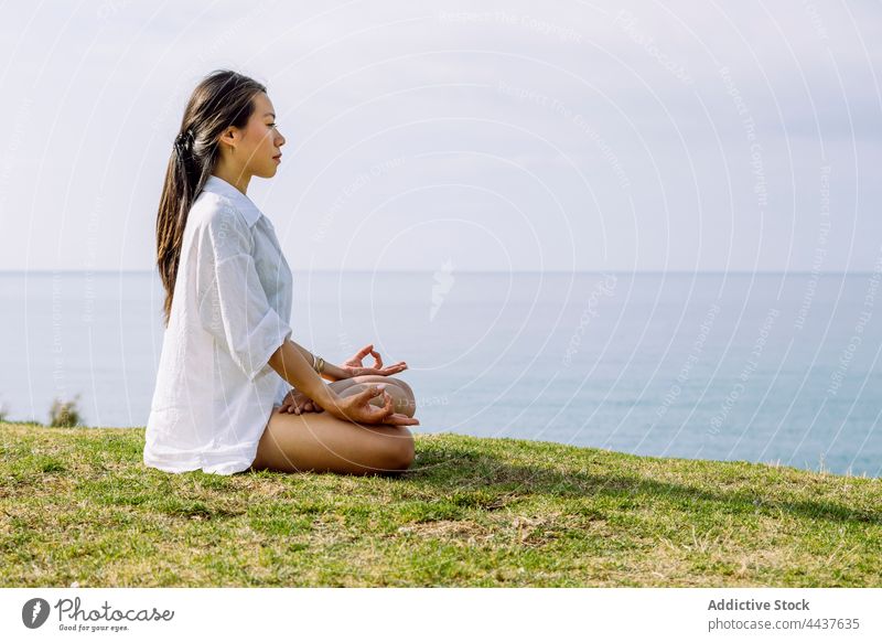 Achtsame asiatische Frau meditiert in Lotus-Pose am grünen Ufer Meditation Yoga Zen Stressabbau Sprit Achtsamkeit Energie Meeresufer Augen geschlossen Vitalität