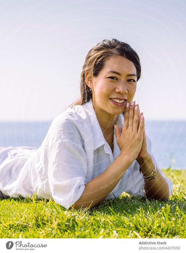 Charmante asiatische Frau mit verschränkten Händen ruht auf Gras Ufer Lächeln Hände gefaltet herzlich charmant freundlich angenehm ruhen Meeresufer Porträt Art